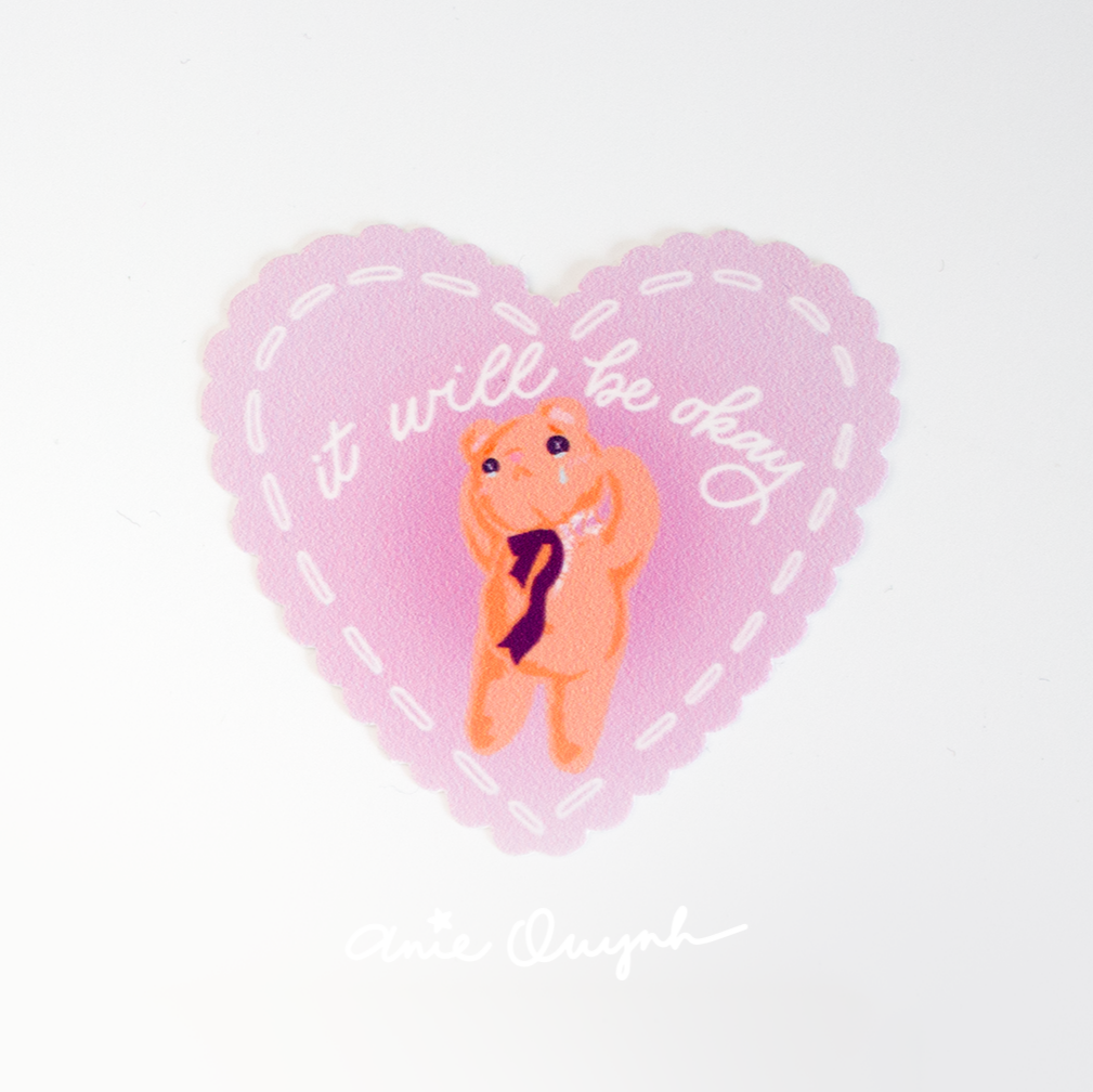 Pink heart sticker with scalloped edges and white "stitching" around the perimeter. In a UV raised gloss coating, it reads "It will be okay" over a drawing of a teddy bear who's crying and losing his head with stuffing coming out.