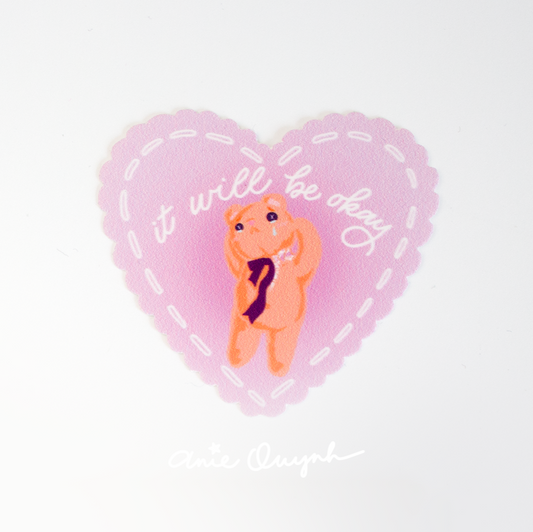 Pink heart sticker with scalloped edges and white "stitching" around the perimeter. In a UV raised gloss coating, it reads "It will be okay" over a drawing of a teddy bear who's crying and losing his head with stuffing coming out.